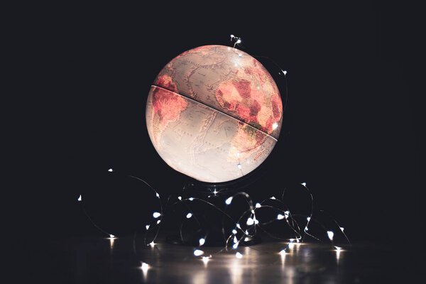 Ascenic  portrait of a light emitting globe stand on a wooden floor in a dark room with lights around it. It depicts earth and is perfect for exploring the world or as a nightstand.