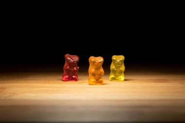 A portrait of three different colored gummi bears, the candy is standing on a wooden plank with lighting that looks like they are on a stage. clipart