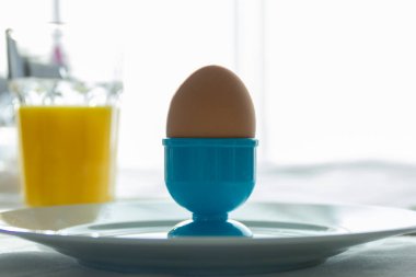 Kahvaltı sırasında, arka planda bir bardak taze portakal suyuyla birlikte, tabakta mavi bir plastik yumurta kabı içinde bir yumurta..