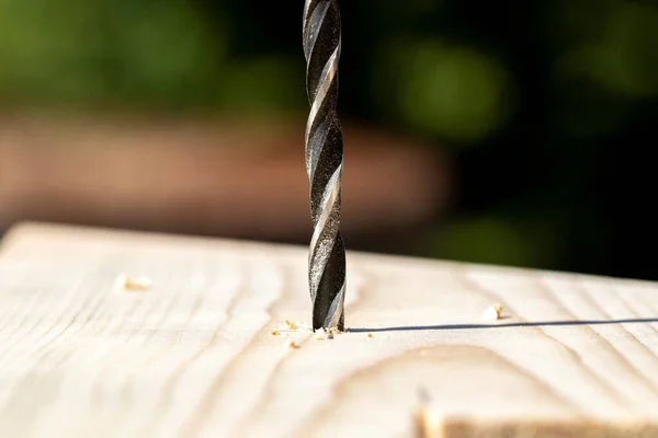 A closeup portrait of a wood drill bit drilling into a wooden plank, with wood chips lying on the work piece.