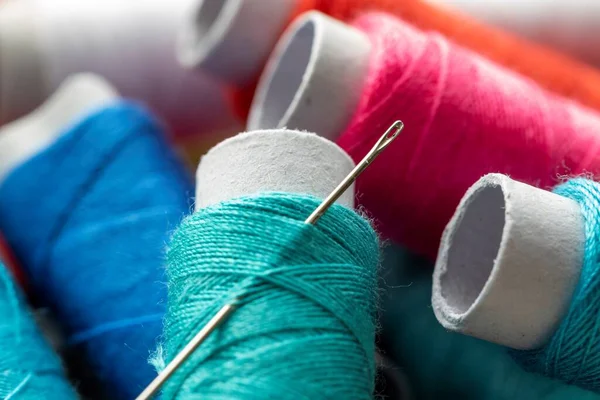 A portrait of a sewing needle stuck in a roll of thread. The spool of wire is lying on top of others rolls, all of them have a different color. The yarn or fiber is ready to be used in soem needlework