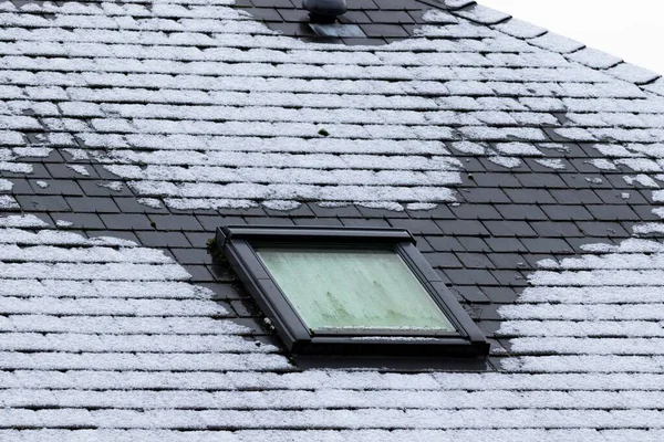 A vertical portrait of a frozen slate roof with a skylight window which is also frozen during winter time. At the sides of the tiltable roof window the frost has molten because of bad isolation.