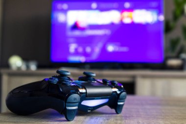 Playstation 4 oyun kontrolörünün Playstation ev menüsünün sergilendiği bir televizyon karşısında yakın plan portresi. PS4 denetleyici çalıştırıldı ve çalıştırılmaya hazır.