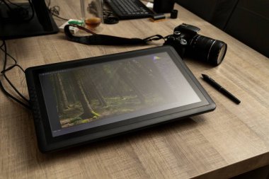 Grafiksel bir kalem tabletinin portresi ve fotoğraf makinesinin yanındaki ahşap masada duran stili. Tablette bir çeşit fotoğraf düzenleme yazılımı var..
