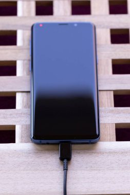 Brecht, Belçika - Nisan 12020: Ahşap bir kutunun üzerinde duran siyah ve mavi bir akıllı telefonun portresi ve limana bağlı bir USB kablosu, pilini şarj ediyor. Şarj ışığı hala kırmızı..