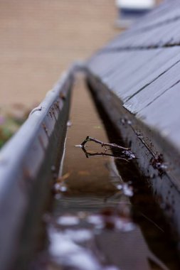 Yağmurlu ve bulutlu bir günde yağmur suyu dolu tıkanmış bir çatı oluğunun portresi. Kanalizasyonun dibi yapraklar ve diğer doğal atıklarla dolu ve tahta bir çatının yanında asılı..
