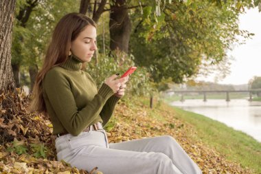 Nehrin kenarında oturan ve boş zamanlarında sıcak bir sonbahar gününde telefona bakan gülümseyen kız. Sonbahar