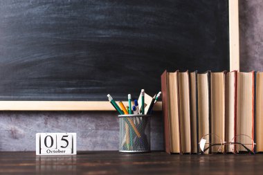 Concept to teacher's day. Pens, pencils, books and glasses on table, on chalkboard background. Calendar october 5, copy space.