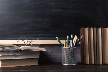 Concept to teacher's day. Pens, pencils, books and glasses on the table, against the background of a chalkboard. Back to school.
