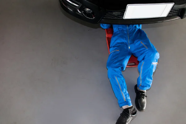 Mechanik v modré uniformě vleže a pracující pod auto — Stock fotografie