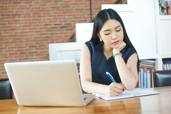 Mulher asiática bonita escrevendo um caderno na mesa com laptop de lado — Fotografia de Stock