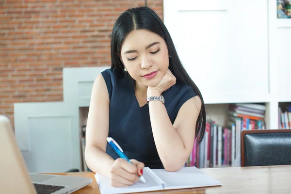 Mulher asiática bonita escrevendo um caderno na mesa com laptop de lado — Fotografia de Stock