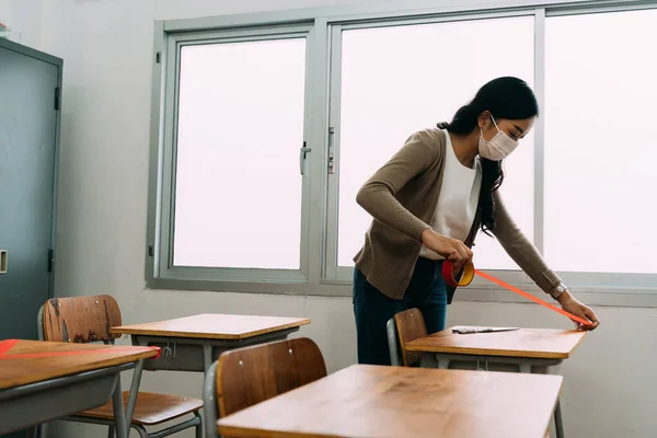 Asiática professora vestindo uma máscara facial mede distâncias entre mesas em sala de aula na escola primária. — Fotografia de Stock