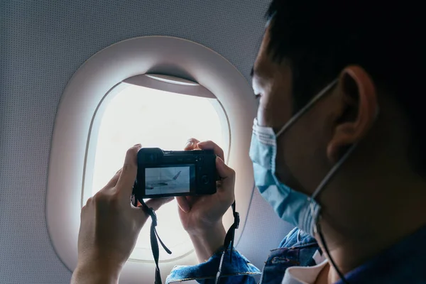 20-talet unga vuxna asiatiska manliga turist bär en ansiktsmask inuti flygplan kabin luft och ta bilder med en kamera. Turism under Covid-19 eller Coronavirus pandemi. — Stockfoto