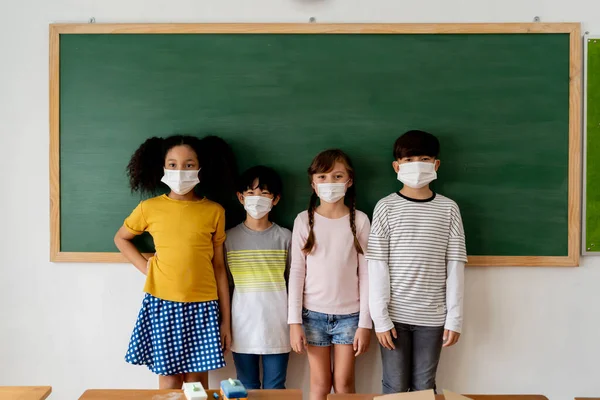 Grupo de diversos estudiantes de primaria en el aula. Alumnos de escuela primaria de pie en fila con una máscara facial con pizarra en el fondo. —  Fotos de Stock