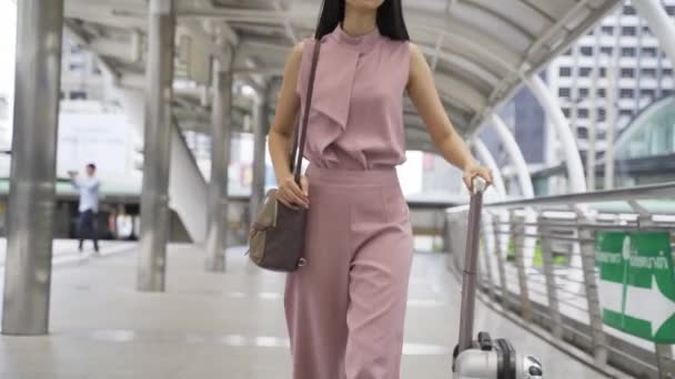 Νεαρή Ασιάτισσα επιχειρηματίας περπατά ενώ σέρνει μια τσάντα τρόλεϊ σε σύγχρονα αστικά κτίρια. Γυναίκα μετακινούμενος πηγαίνει για επαγγελματικό ταξίδι. — Αρχείο Βίντεο
