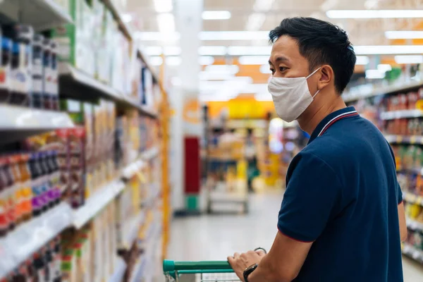 Young adult Asian man wearing a face mask while shopping with cart trolley in grocery supermarket store.