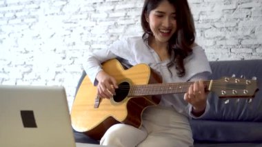 Genç Asyalı kadın dizüstü bilgisayar monitöründen gitar çalmayı öğreniyor. Kadın gitarist çevrimiçi özel ders izliyor.