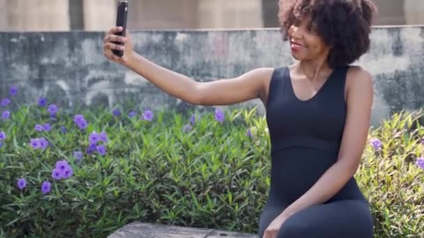 Ung afroamerikansk kvinna som använder en mobiltelefon och tar selfie foto medan du sitter på en bänk i parken. Kvinnlig svart joggare vilar och använder en smartphone. — Stockvideo