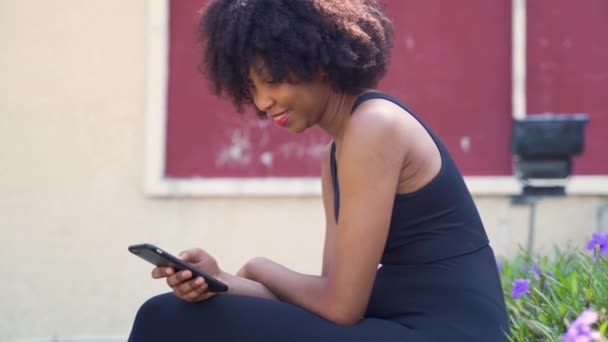 Genç Afrikalı Amerikalı kadın cep telefonu kullanıyor ve parkta bir bankta oturuyor. Kadın siyah koşucu dinleniyor ve dışarıda gülümsüyor. — Stok video