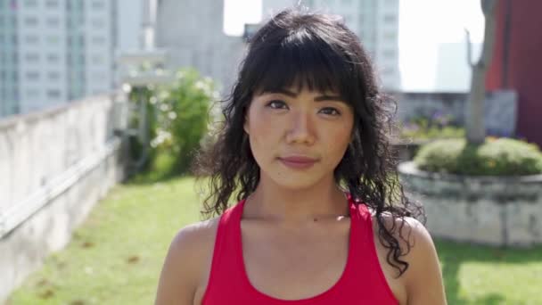 Fiatal ázsiai nő sportruházatban néz a kamerába és mosolyog a kültéri tetőtéri parkban. Boldog és gondtalan női edző nyáron. — Stock videók