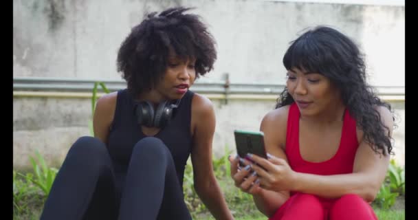 Groupe diversifié de deux amies de fitness parlant et utilisant un téléphone portable dans le parc. Jeunes femmes au repos et souriantes à l'extérieur — Video