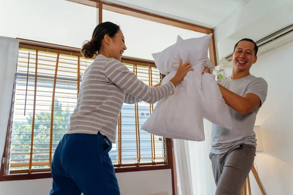 Portrait of young adult Asian couple playing pillow fight in bedroom interior scene. 30s mature husband and wife smiling and having a fun activity. Marriage and happy relationship life concept.