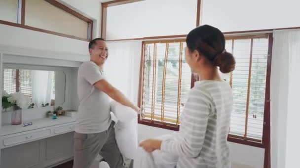 寝室の内部の場面で枕の戦いをしている若い大人のアジアのカップル。30代の成熟した夫と妻が笑顔で楽しい活動をしています。結婚と幸せな関係生活の概念 — ストック動画