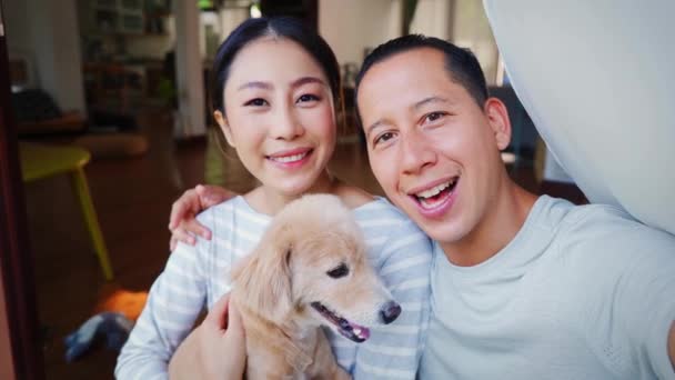 Fiatal ázsiai házaspár kezében egy kiskutya, akik szelfit készítenek egy telefonról, otthon a háttérben. 30-as évekbeli felnőtt férfi és nő kutyával, akik családi fotókat készítenek. - Boldog csoportképet. — Stock videók