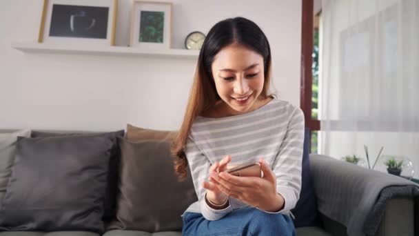 30-talet ung vuxen asiatisk kvinna spelar en mobiltelefon sitter på soffan i vardagsrummet hemma. Glad kvinnlig internet och teknik användare i casual kläder på soffan i mysiga rum — Stockvideo