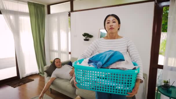 Mladá žena nosí košík špinavých šatů a doma dělá domácí práce, zatímco líný muž odpočívá a spí na pohovce. Pár s domácím problémem domácí práce. — Stock video