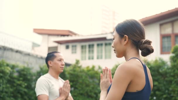 Jong fitnesspaar dat zonnesaluutyoga beoefent poseert buiten in de tuin van het huis. Aziatische vrouw en blanke man training buiten in het park — Stockvideo