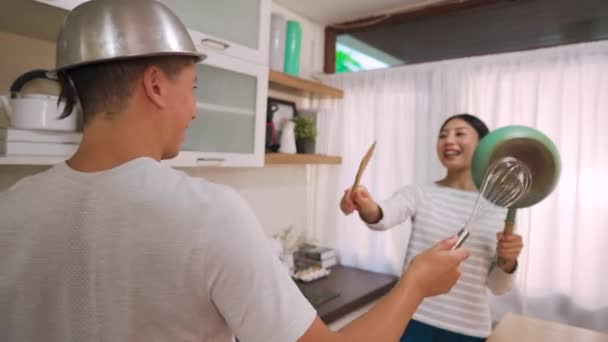คู่สมรสหนุ่มสาวกําลังทําตัวเป็นคนธรรมดาต่อสู้กับเครื่องใช้และกระทะในครัวที่บ้าน สามีและภรรยาที่ตลกถือเครื่องครัวมีช่วงเวลาที่สนุก — วีดีโอสต็อก
