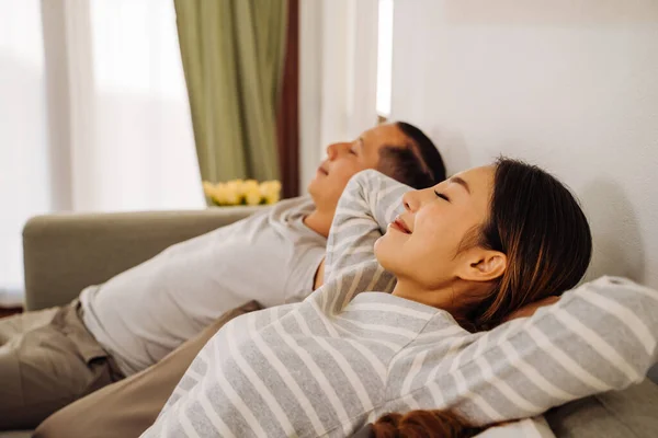 Vista laterale di una coppia che si rilassa su un divano in soggiorno a casa. caucasico e asiatico donna respirazione e riposo mentre sorridente e sdraiato in divano indoor — Foto Stock