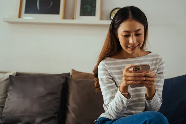 30s young adult Asian woman playing a mobile phone sits on sofa in the living room at home. Happy female internet and technology user in casual clothes on couch in cozy room. — Stock Photo, Image