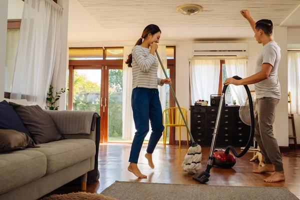 Happy young couple of woman dancing and singing while cleaning with vacuum cleaner machine and man mopping the floor in living room at home