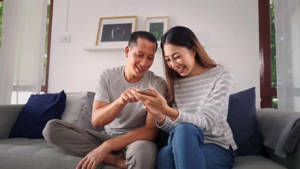 In den 30er Jahren sitzen junge erwachsene asiatische Männer und Frauen, die zusammen ein Mobiltelefon spielen, zu Hause auf dem Sofa im gemütlichen Wohnzimmer. Glückliches Paar Internetnutzer in lässiger Kleidung auf der Couch. Technologieeinsatz im Asien-Konzept — Stockvideo