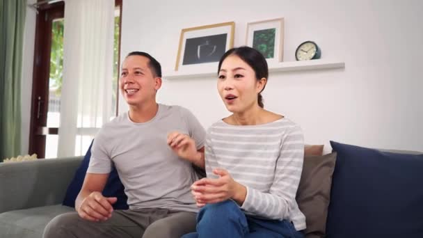 Portrait of 30s young adult husband and wife sitting on couch at home and watching entertainment on tv. Asian happy and joyful couple enjoy leisure time and laughing in cozy living toom together. — Stock Video