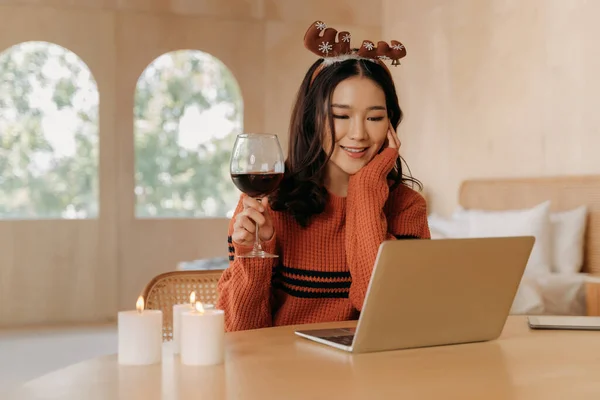 若い大人のアジアの女性は、テーブルの上にワインとキャンドルのガラスと屋内部屋でトーストを作るセーターとトナカイの耳を身に着けています。クリスマスの祭りの間にオンラインでラップトップビデオ通話を使用してかわいい女の子. — ストック写真