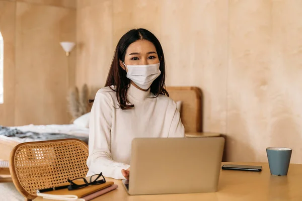 아시아의 젊은 여성은 인터넷에서 집에서 컴퓨터로 일하는 컴퓨터 랩탑을 사용하여 스웨터를 입는다. 코비 디온 -19 의 확산을 막기 위해 마스크를 쓴 매력적 인 소녀 - 코로나 바이러스 유행. 복사 공간. — 스톡 사진
