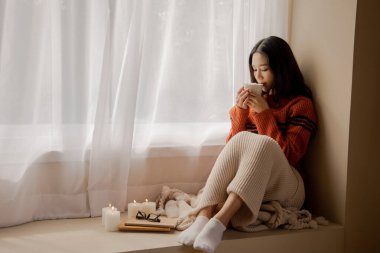 Young beautiful Asian woman holding cup of coffee and marshmallows on top, sitting at home and looking out the window. Happy girl drinking chocolate in sweater in cold winter clipart