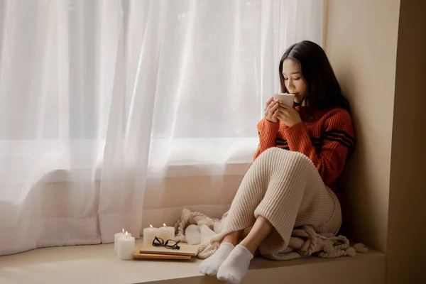Молодая красивая азиатка, держащая чашку кофе и зефир сверху, сидит дома и смотрит в окно. Счастливая девушка пьет шоколад в свитере холодной зимой — стоковое фото