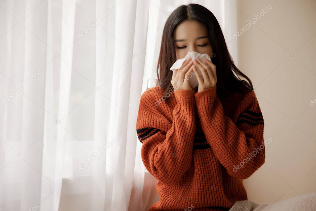 Young Asian ill woman with handkerchief coughing and sneezing, near window at home. Sick girl holding a tissue napkin staying at home during Coronavirus Covid 19 pandemic isolation