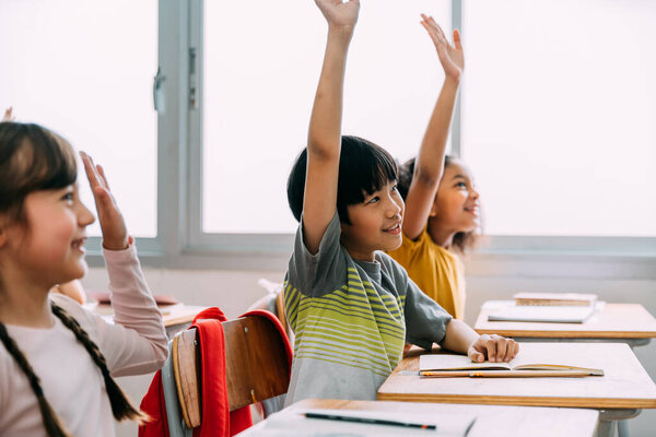 Elementary age Asian student boy raised hands up in Q and A class. Diverse group of pre-school pupils in elementary age in education building school. Volunteering and participating classroom concept