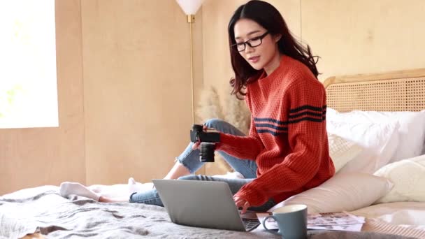 Ung asiatisk kvinnlig fotograf i tröja arbetar på laptop på sängen. Glad kvinna revidera bilder tagna i datorn anteckningsbok. Vinterporträtt mode — Stockvideo