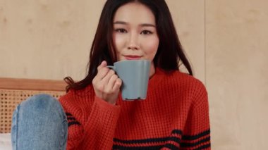 Young beautiful Asian woman looking at camera and holding cup of coffee and marshmallows on top, sitting at home . Happy girl drinking chocolate in sweater in cold winter