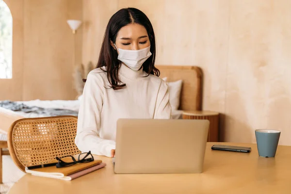 Jovem adolescente mulher asiática usa suéter usando laptop computador trabalhando em casa dentro de casa online. Menina atraente usando uma máscara facial para evitar a propagação de Covid-19 - pandemia de Coronavirus. Espaço de cópia. — Fotografia de Stock