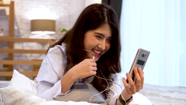 Retrato de menina asiática feliz em roupas casuais deitado na cama enquanto faz uma chamada de vídeo com smartphone e acenando no quarto em casa. Conceito de tecnologia de videoconferência — Vídeo de Stock