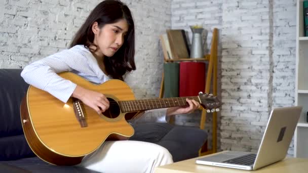 Mujer asiática joven practicando y aprendiendo a tocar la guitarra en el monitor del ordenador portátil. Guitarrista femenina viendo tutorial en línea — Vídeos de Stock