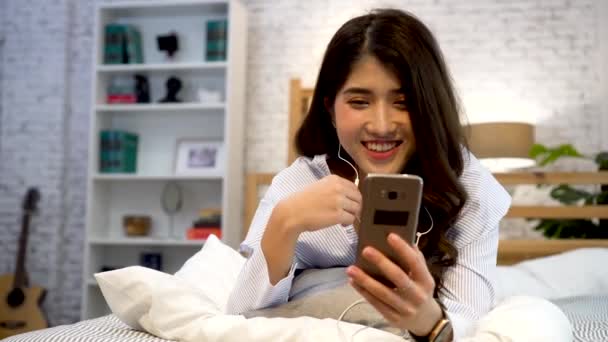 Porträt eines glücklichen jungen asiatischen Mädchens in lässiger Kleidung, das auf dem Bett liegt, während es ein Videotelefon mit dem Smartphone macht und im Schlafzimmer zu Hause winkt. Videokonferenztechnologiekonzept — Stockvideo